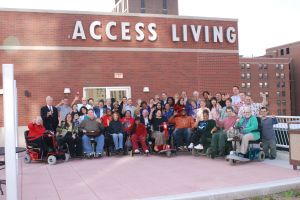 a photo of access living board and staff. the photo is taken on the roof of Access Living's building in Chicago. The group of people is standing against a brick wall, on which, in large block letters, is the name "Access Living"