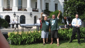 two men, both wearing shorts and both in green tshirts, stand on the White House South Lawn.  They stand side by side with outside arms raised up and forward, with fists clenched in a show of solidarity and power. 