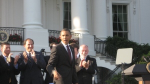 In this photo, President Obama is approaching the podium on the South Lawn of the White House.  Several men on in the background, applauding his approach, including, on the far right, Robert David Hall, who introduced the president. 